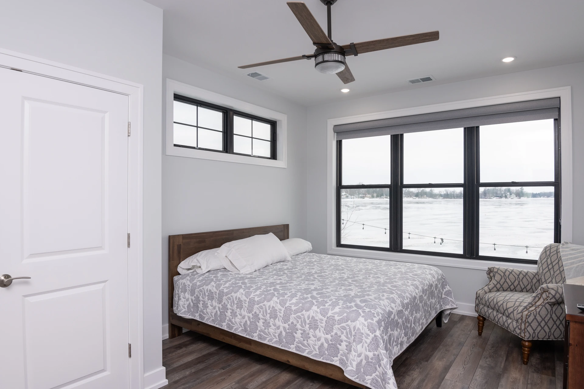 Bedroom with black-framed lake view windows, wood bed frame, patterned bedding, ceiling fan, and dark wood-look flooring