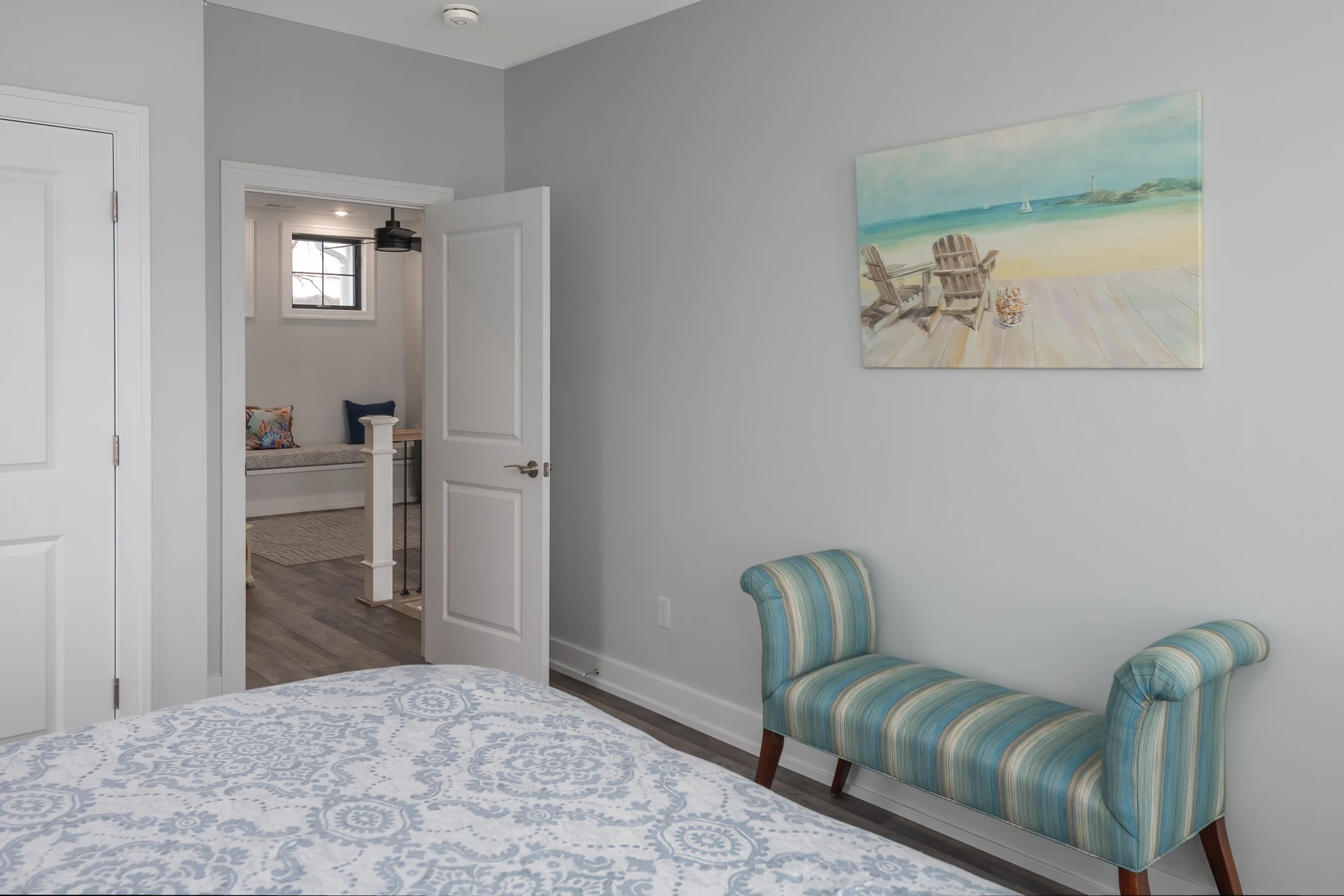 Bedroom with gray walls, patterned bedding, striped accent bench, beach artwork, and doorway to adjoining sitting area