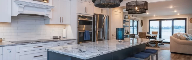 Blue Island Kitchen with Open Living View | Pennings & Sons Open kitchen with blue island, white cabinetry, marble-look countertops, bar stools, and lakefront living area beyond