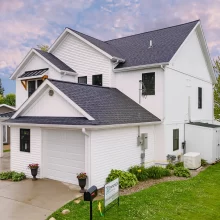 Side exterior of white lakefront home with attached garage, dark shingle roof, landscaped foundation, and lake visible beyond