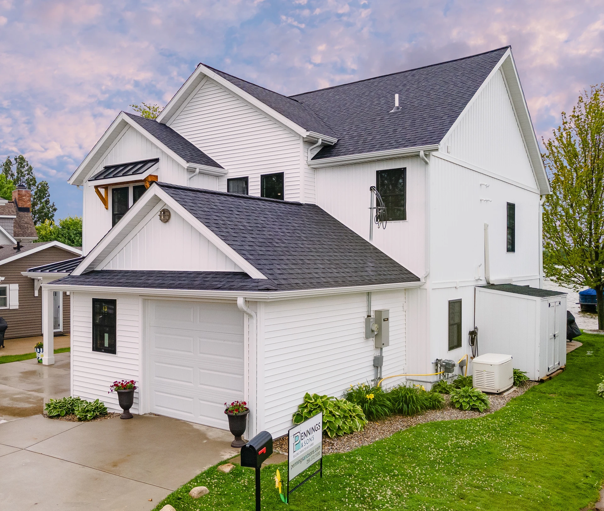 Side exterior of white lakefront home with attached garage, dark shingle roof, landscaped foundation, and lake visible beyond