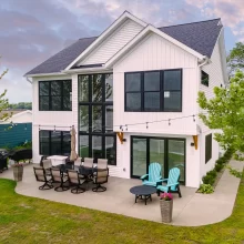 White lakefront home with tall black-framed windows, rear patio, outdoor seating, and lawn leading toward the water