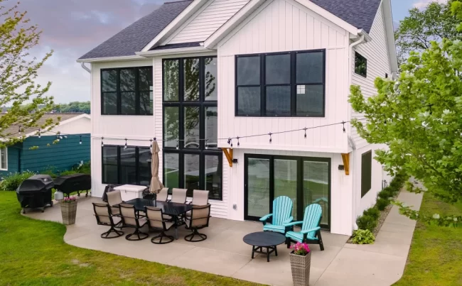 White Lakefront Home Rear Exterior | Pennings & Sons White lakefront home with tall black-framed windows, rear patio, outdoor seating, and lawn leading toward the water