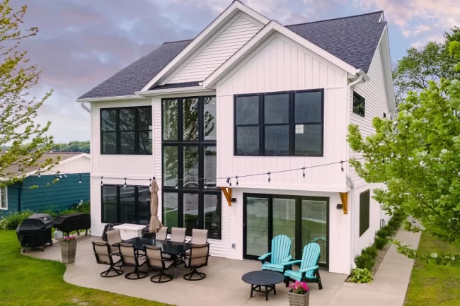 White Lakefront Home Rear Exterior | Pennings & Sons White lakefront home with tall black-framed windows, rear patio, outdoor seating, and lawn leading toward the water