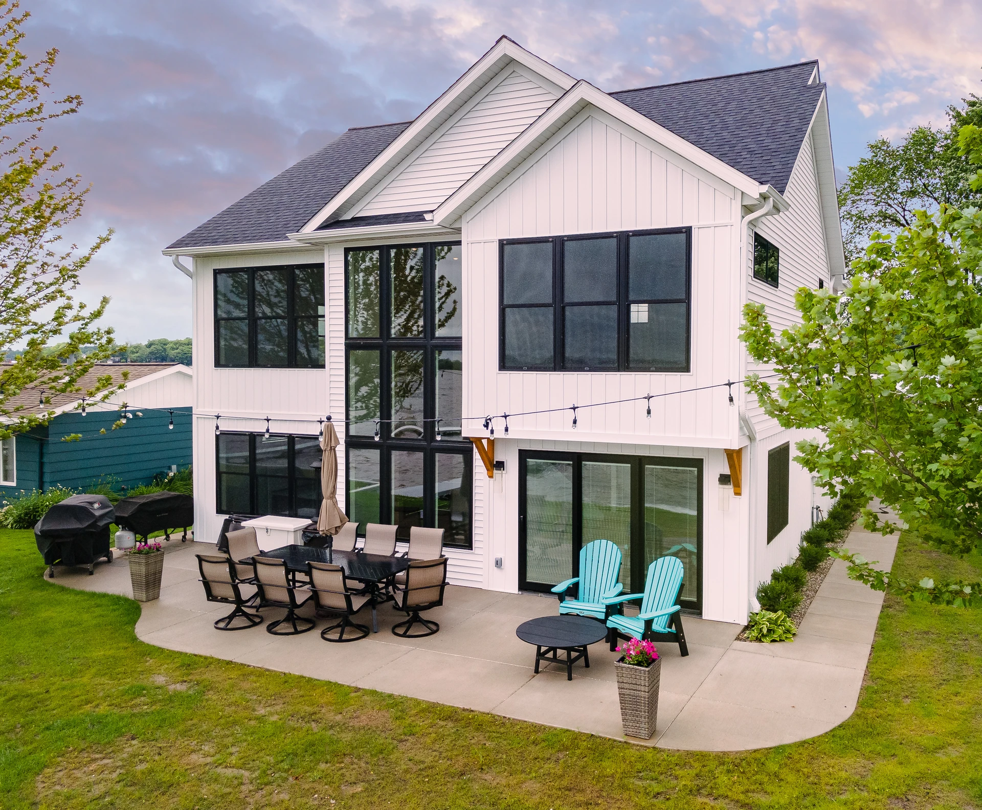 White lakefront home with tall black-framed windows, rear patio, outdoor seating, and lawn leading toward the water