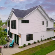 White lakefront home exterior with black windows, side patio seating, concrete walkway, and lake views behind the house