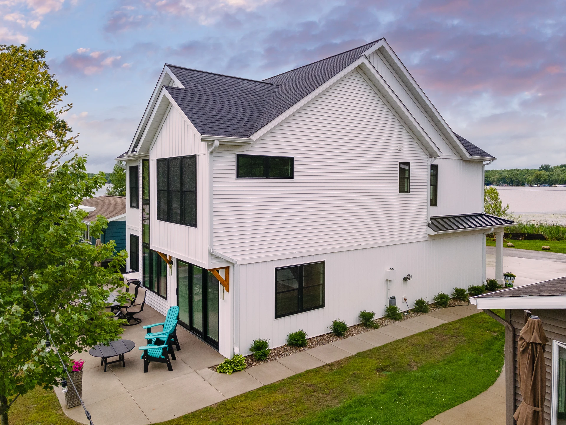 White lakefront home exterior with black windows, side patio seating, concrete walkway, and lake views behind the house