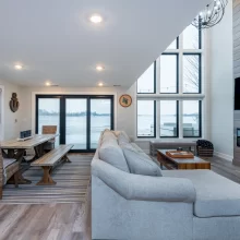 Lakefront living room with tall black-framed windows, dining area, sectional sofa, and wood fireplace feature wall