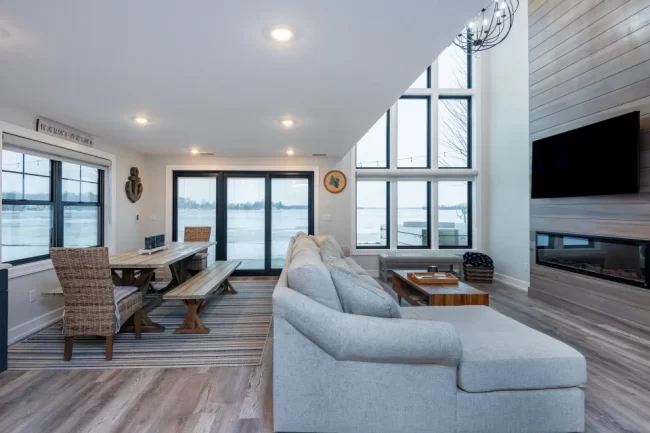 Lakefront Living Room with Tall Windows | Pennings & Sons Lakefront living room with tall black-framed windows, dining area, sectional sofa, and wood fireplace feature wall