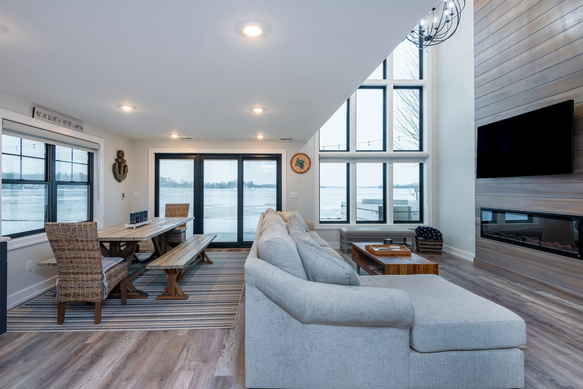 Lakefront living room with tall black-framed windows, dining area, sectional sofa, and wood fireplace feature wall