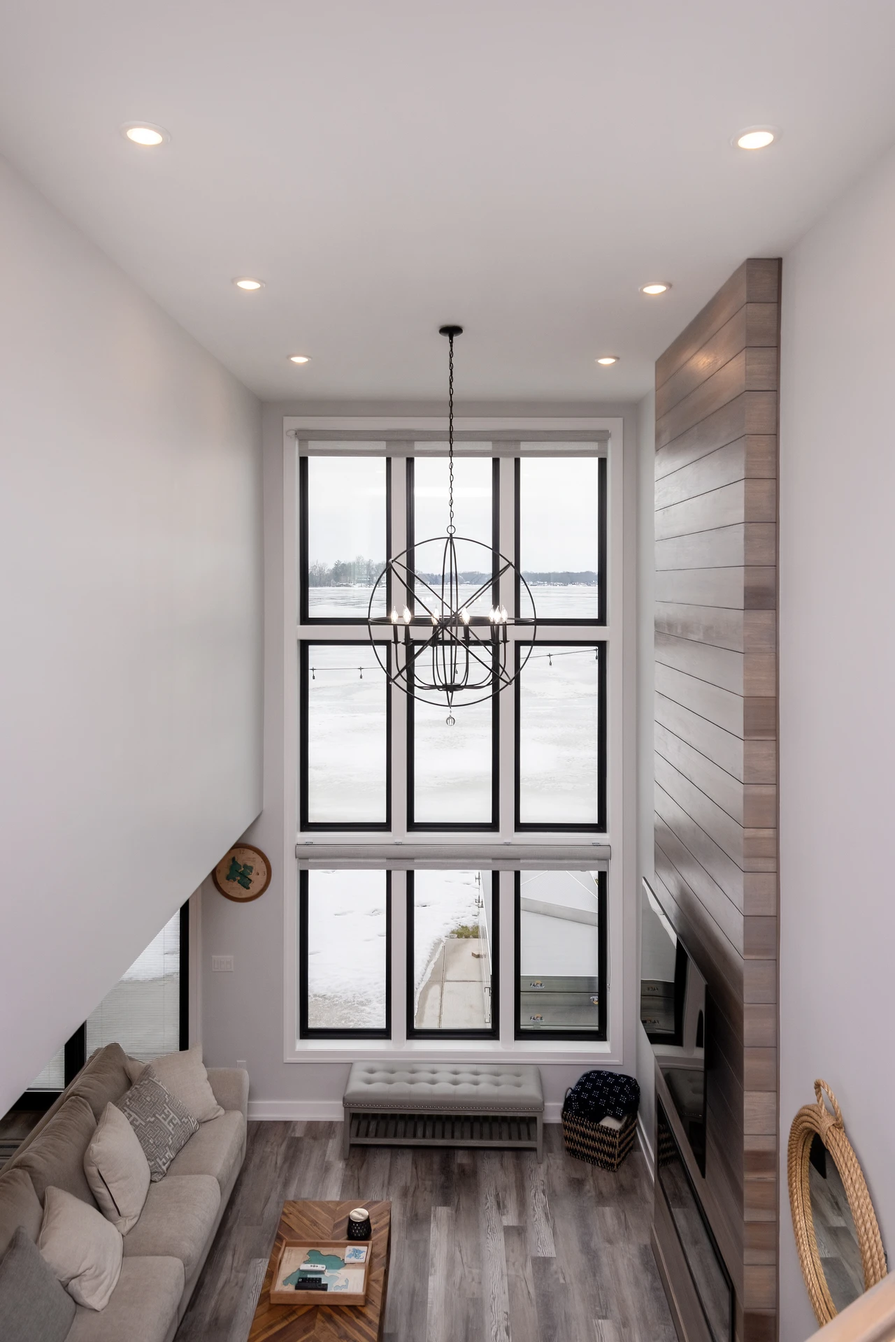 Overhead view of two-story living room with large lake-facing window, chandelier, and tall wood fireplace wall