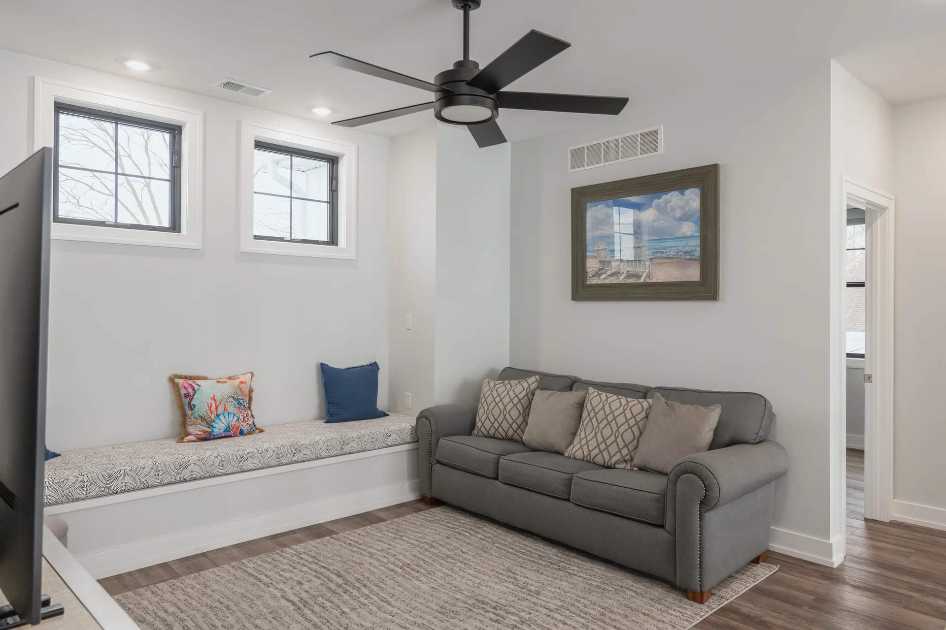 Loft Sitting room with gray sofa, built-in window bench, black-framed windows, ceiling fan, and coastal wall art
