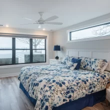 Bedroom with lake view window, blue floral bedding, white headboard, ceiling fan, and gray wood-look flooring