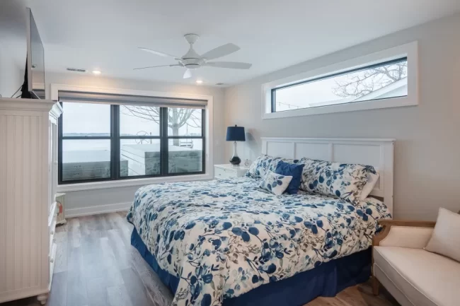 Lake View Bedroom with Blue Floral Bedding | Pennings & Sons Bedroom with lake view window, blue floral bedding, white headboard, ceiling fan, and gray wood-look flooring