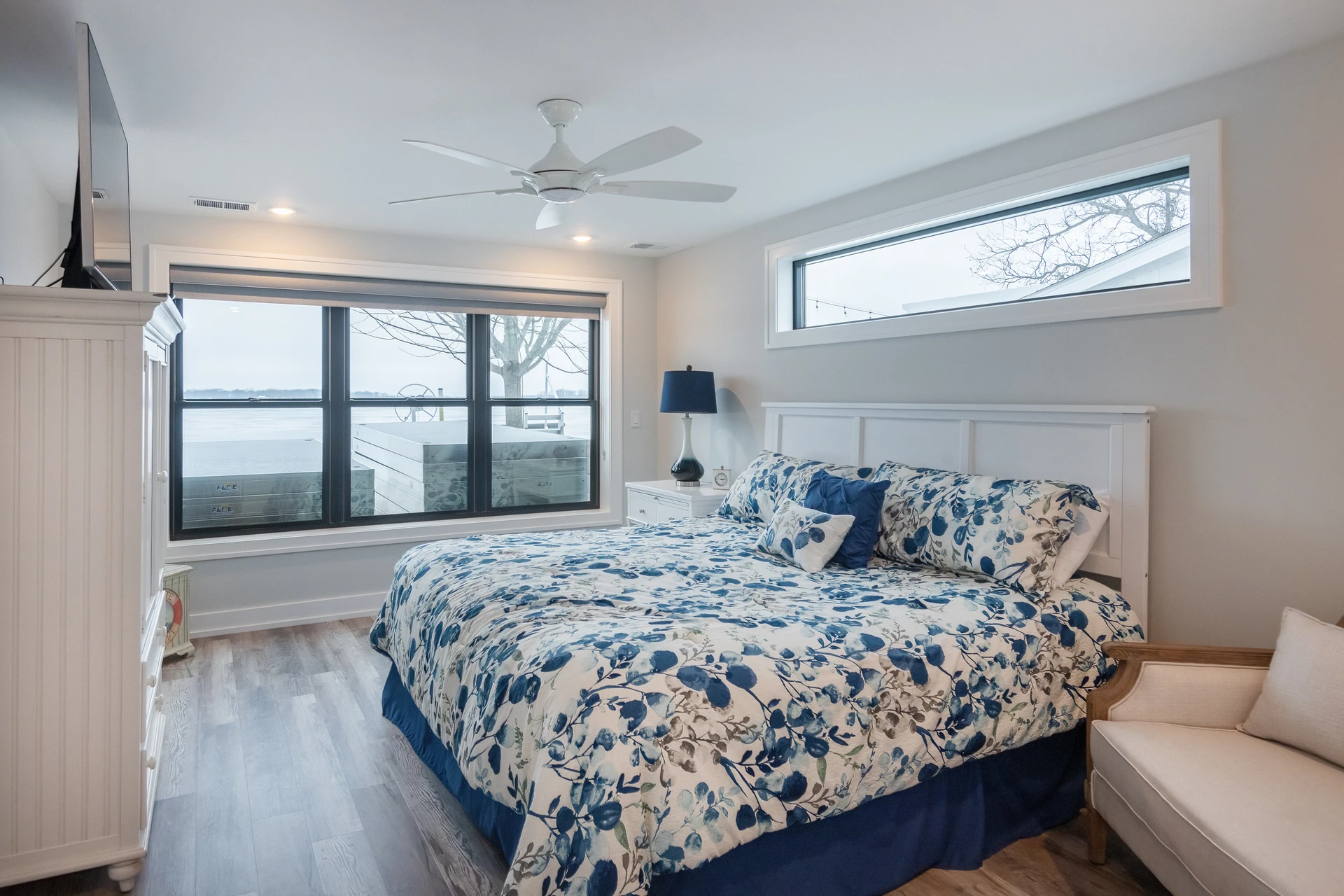 Bedroom with lake view window, blue floral bedding, white headboard, ceiling fan, and gray wood-look flooring