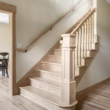 Natural wood staircase with custom newel post, matching railing, light wood flooring, and open foyer detail