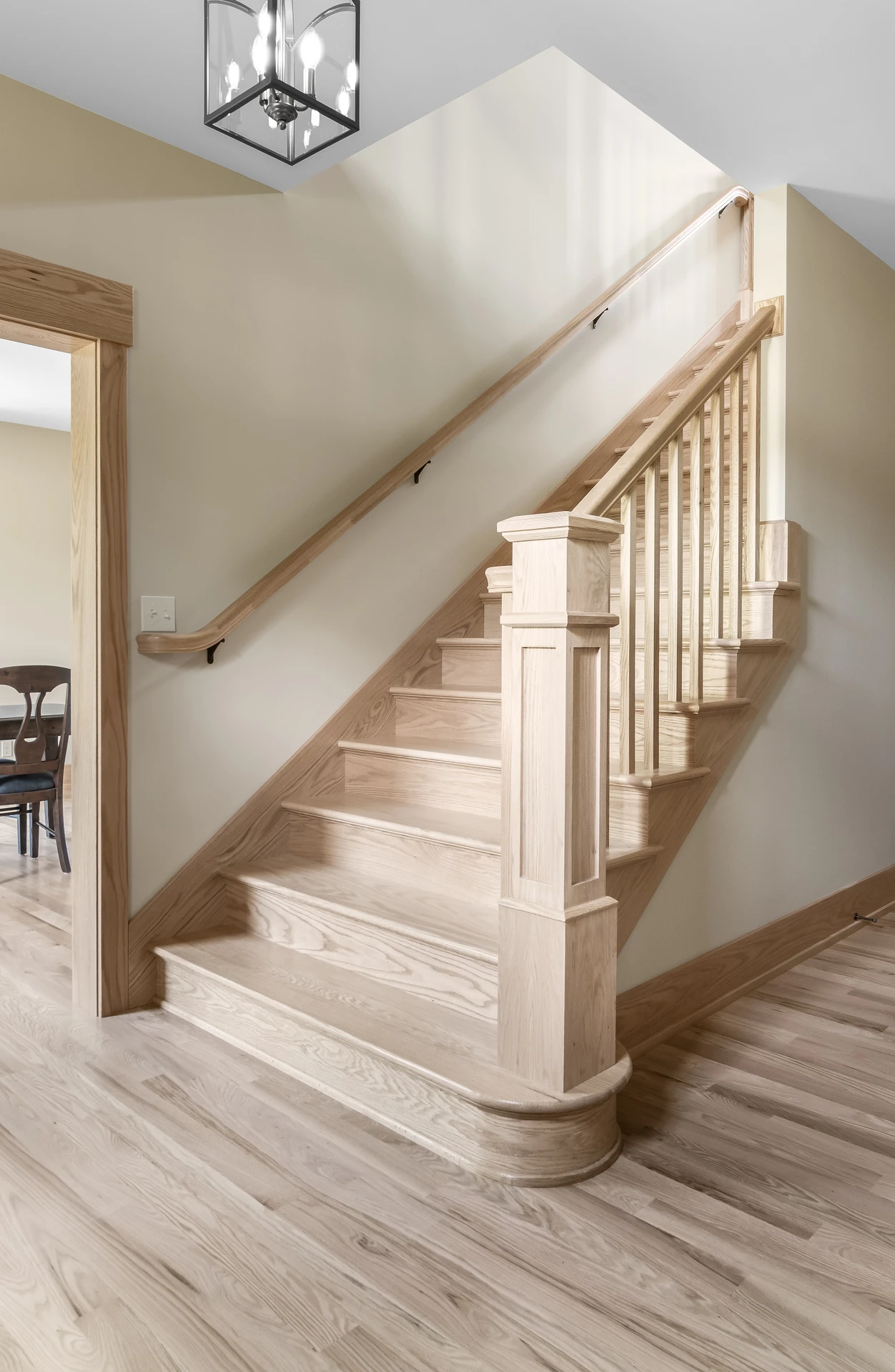 Natural wood staircase with custom newel post, matching railing, light wood flooring, and open foyer detail