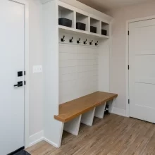 Built-in mudroom bench with white shiplap backing, black coat hooks, upper storage cubbies, wood bench seat, and entry door