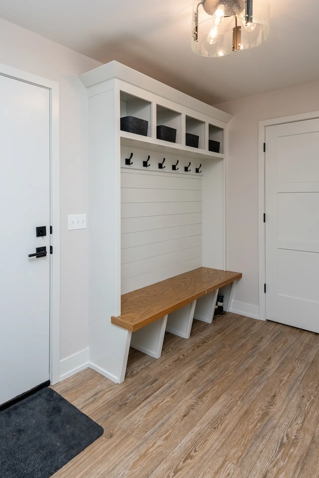 Built-in mudroom bench with white shiplap backing, black coat hooks, upper storage cubbies, wood bench seat, and entry door