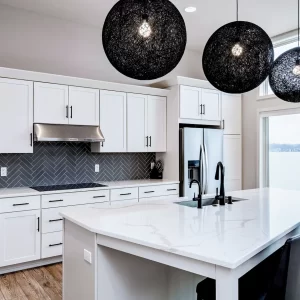 Custom white kitchen with large quartz island, black bar stools, stainless refrigerator, gray herringbone backsplash, and black fixtures