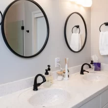 Double bathroom vanity with white speckled countertop, black faucets, round black mirrors, towel rings, and white cabinetry