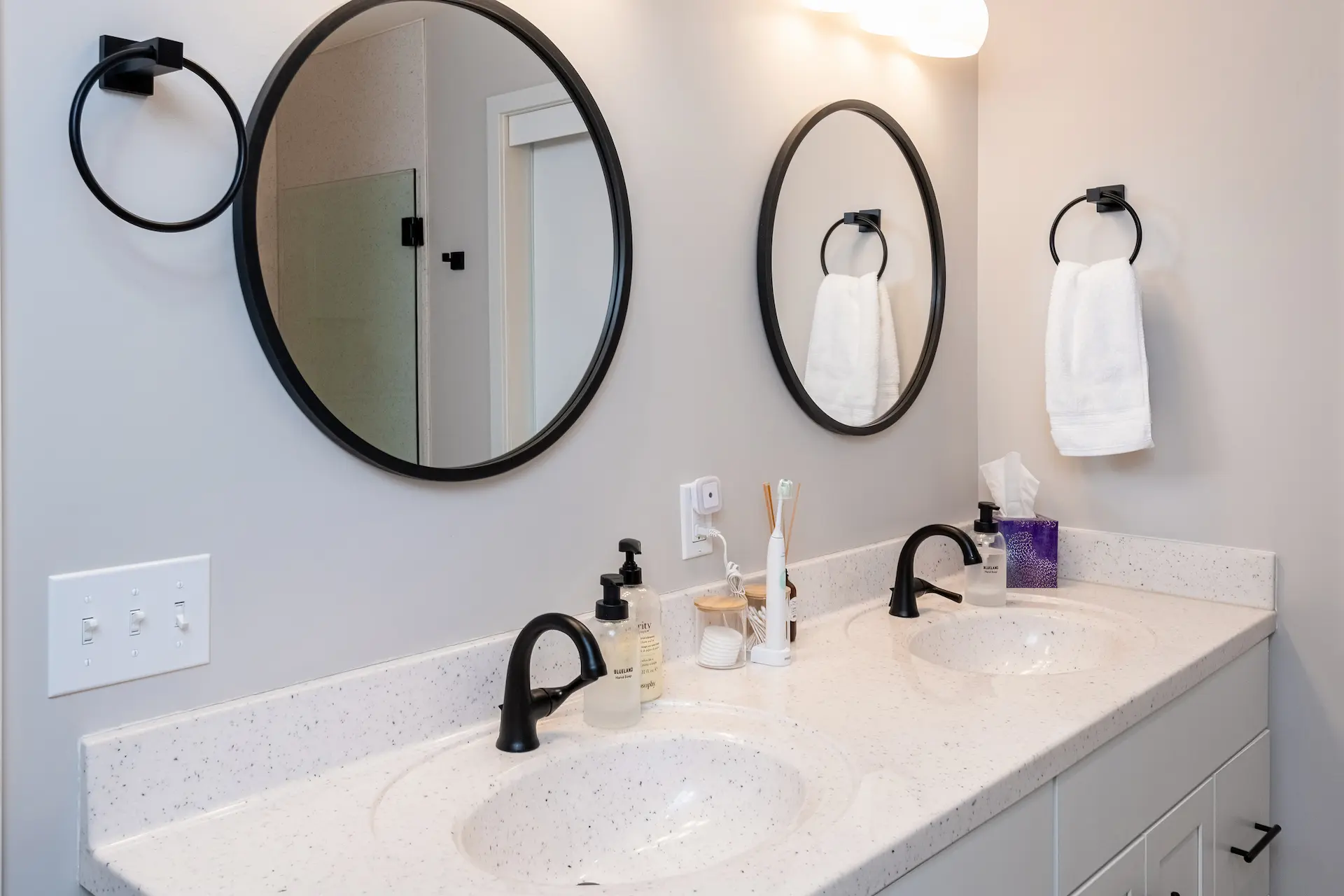 Double bathroom vanity with white speckled countertop, black faucets, round black mirrors, towel rings, and white cabinetry