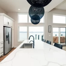 Large white kitchen island with black pendant globes, black faucet, stainless refrigerator, and wide lake-facing windows