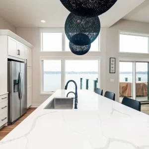 Large white kitchen island with black pendant globes, black faucet, stainless refrigerator, and wide lake-facing windows