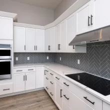 White kitchen range wall with gray herringbone tile backsplash, stainless steel hood, built-in double ovens, quartz counters, and black hardware
