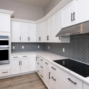 White kitchen range wall with gray herringbone tile backsplash, stainless steel hood, built-in double ovens, quartz counters, and black hardware