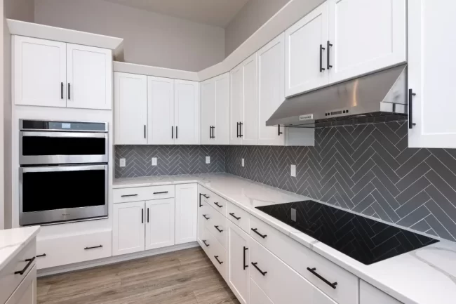 White kitchen range wall with gray herringbone tile backsplash, stainless steel hood, built-in double ovens, quartz counters, and black hardware