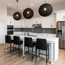 White kitchen with quartz island, black bar stools, black globe pendant lights, stainless appliances, and gray herringbone tile backsplash