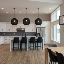 White kitchen with large island, black bar stools, gray herringbone backsplash, black globe pendant lights, and lake-facing windows