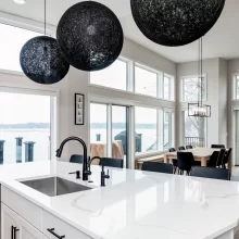 Lakefront kitchen with large white island, black globe pendants, sink, dining table, sliding doors, and waterfront views