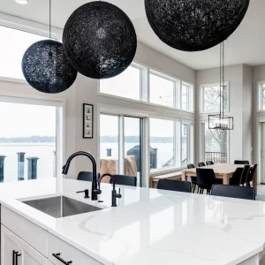 Lakefront kitchen with large white island, black globe pendants, sink, dining table, sliding doors, and waterfront views