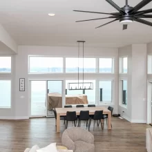 Open concept dining room with large lake-facing windows, wood table, black chairs, modern chandelier, kitchen island, and vaulted ceiling fan