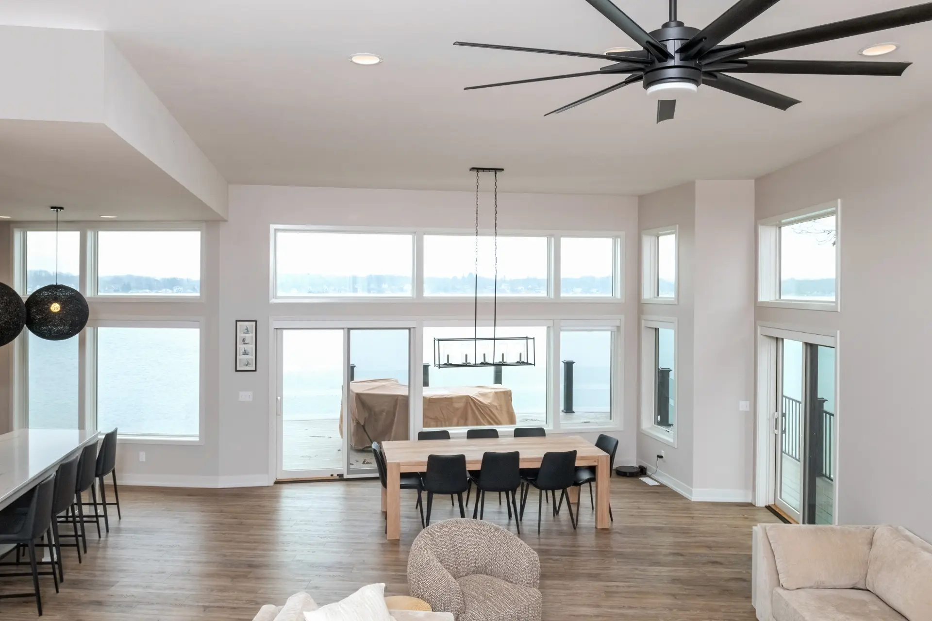 Open concept dining room with large lake-facing windows, wood table, black chairs, modern chandelier, kitchen island, and vaulted ceiling fan