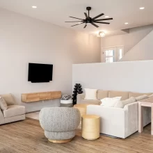Modern living room with beige sectional, wall-mounted TV, wood flooring, black ceiling fan, and staircase with white half wall