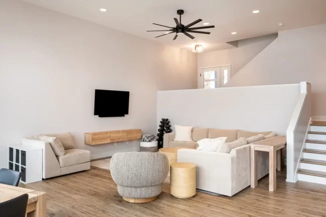 Modern Living Room with Staircase | Pennings & Sons Modern living room with beige sectional, wall-mounted TV, wood flooring, black ceiling fan, and staircase with white half wall