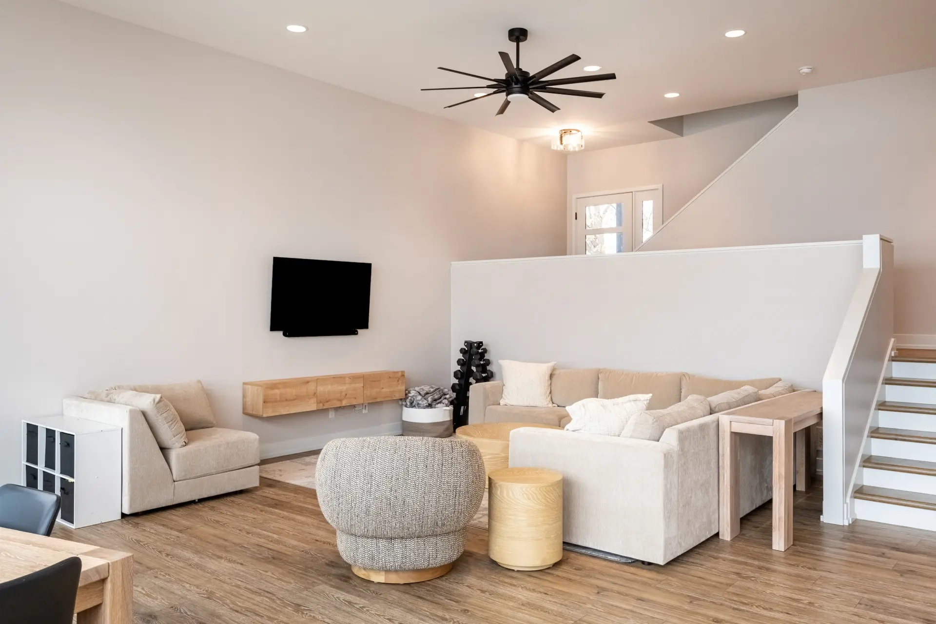 Modern living room with beige sectional, wall-mounted TV, wood flooring, black ceiling fan, and staircase with white half wall