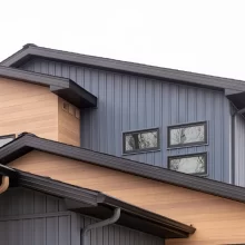Modern home exterior detail with gray vertical siding, warm wood-look accent siding, black roof trim, gutters, and upper-level windows