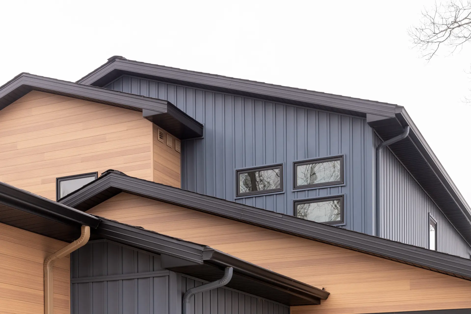 Modern home exterior detail with gray vertical siding, warm wood-look accent siding, black roof trim, gutters, and upper-level windows
