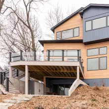 Rear exterior of a modern lakefront home with warm wood siding, dark vertical siding, upper deck, glass railing, stairs, and wooded yard