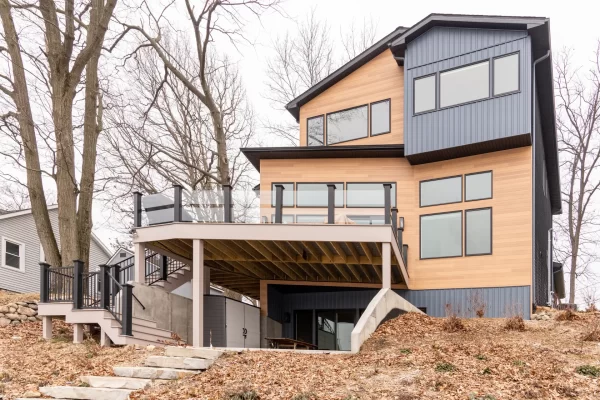 Rear exterior of a modern lakefront home with warm wood siding, dark vertical siding, upper deck, glass railing, stairs, and wooded yard