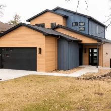 Modern lakefront home exterior with wood-look siding, black garage door, covered entry, dark trim, lakeside railing, and concrete driveway