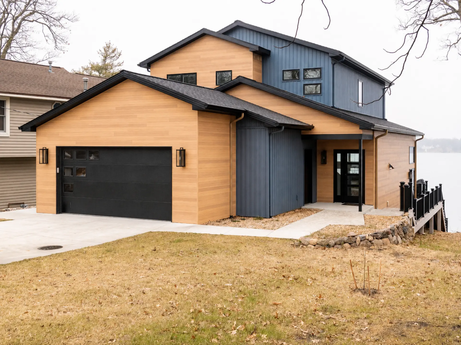 Modern lakefront home exterior with wood-look siding, black garage door, covered entry, dark trim, lakeside railing, and concrete driveway