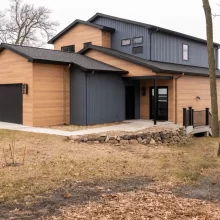 Modern lakefront home exterior with warm wood siding, dark vertical siding, black garage door, stone retaining wall, deck railing, and wooded yard