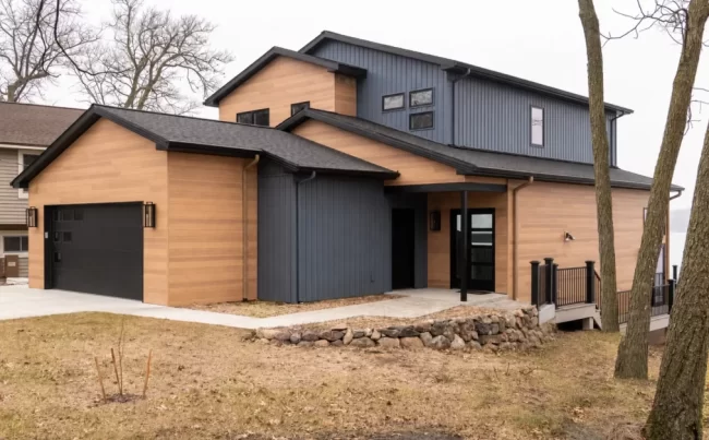 Modern Lakefront Home Exterior Side View | Pennings & Sons Modern lakefront home exterior with warm wood siding, dark vertical siding, black garage door, stone retaining wall, deck railing, and wooded yard