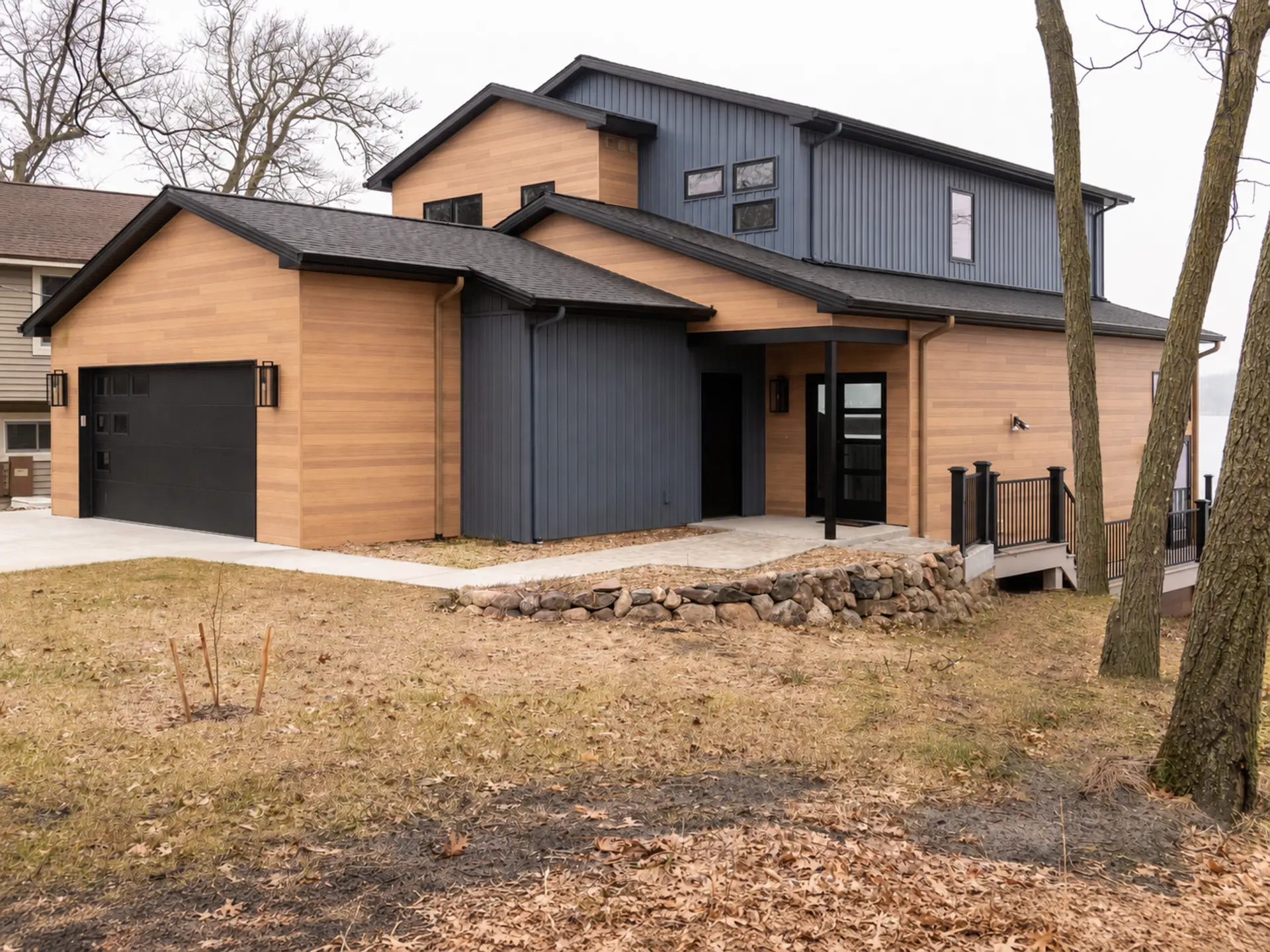 Modern lakefront home exterior with warm wood siding, dark vertical siding, black garage door, stone retaining wall, deck railing, and wooded yard