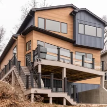 Modern lakefront home exterior with multi-level deck, glass railing, exterior stairs, black railings, wood-look siding, and dark vertical siding